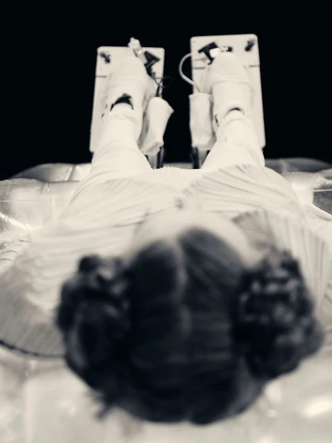 Black and white eye-level shot of a woman lying down with legs elevated on cushioned supports, with dark hair and striped clothing.