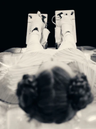 Black and white eye-level shot of a woman lying down with legs elevated on cushioned supports, with dark hair and striped clothing.