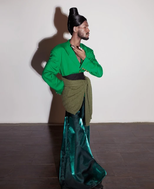 Profile shot of an adult male with a high-fashion hairstyle, green jacket, and emerald skirt against a neutral studio backdrop.