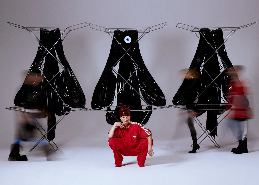 A woman in a red Adidas tracksuit squats in front of three garment racks draped with black fabric, with blurred figures in the background.