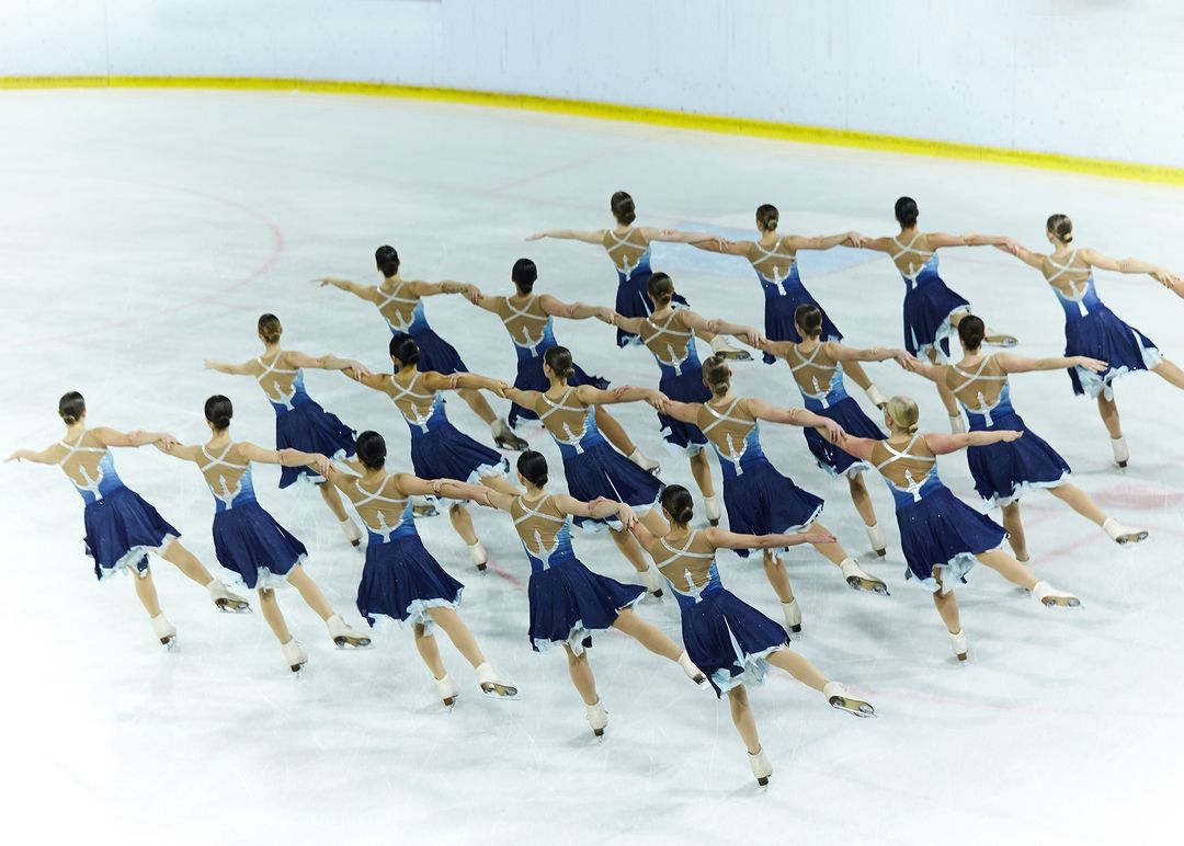 Synchronized ice skating team performs on an ice rink, wearing matching blue dresses with white trim, arms extended outward.