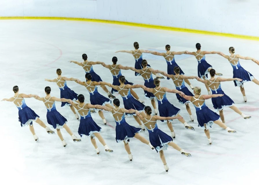 Synchronized ice skating team performs on an ice rink, wearing matching blue dresses with white trim, arms extended outward.