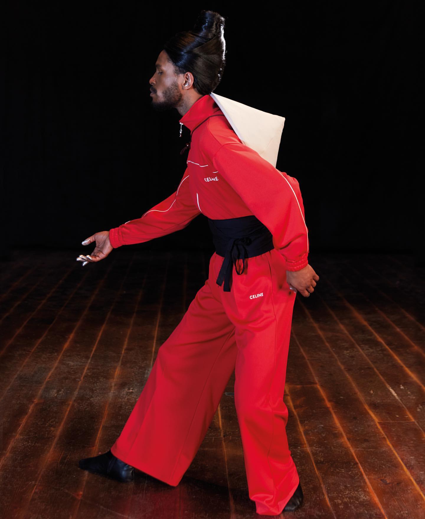 Fashion portrait of a Black male model in a red tracksuit with a black obi belt against a black background and wooden floor.