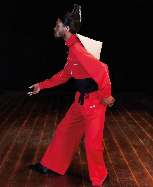 Fashion portrait of a Black male model in a red tracksuit with a black obi belt against a black background and wooden floor.