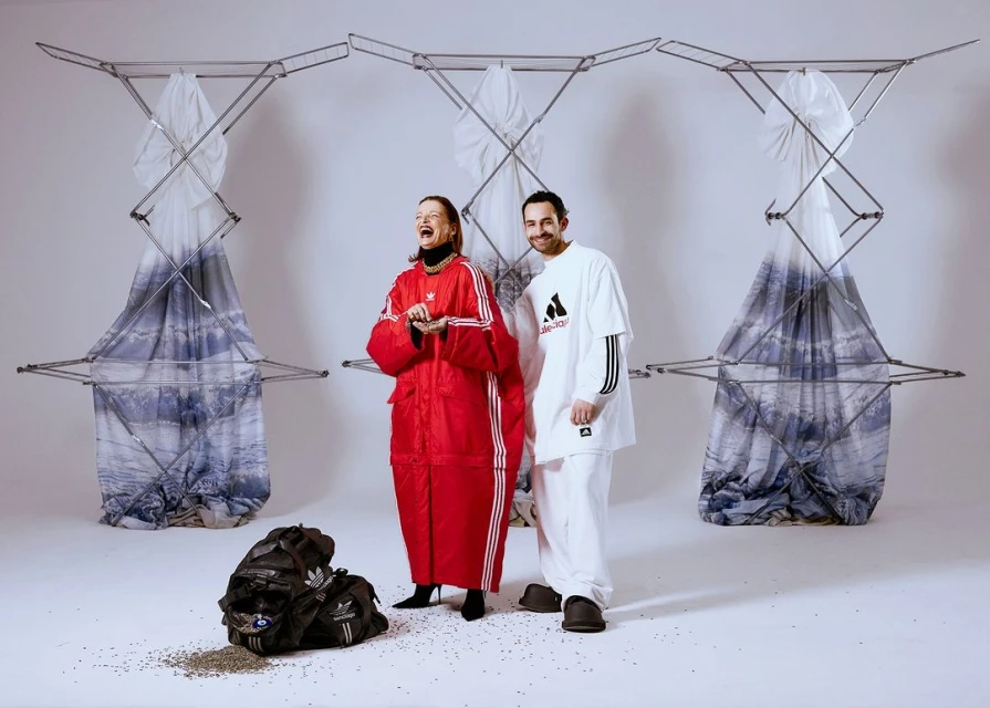 Two adults in Adidas apparel strike a pose amidst abstract clothing racks and scattered seeds in a bright, white studio setting.