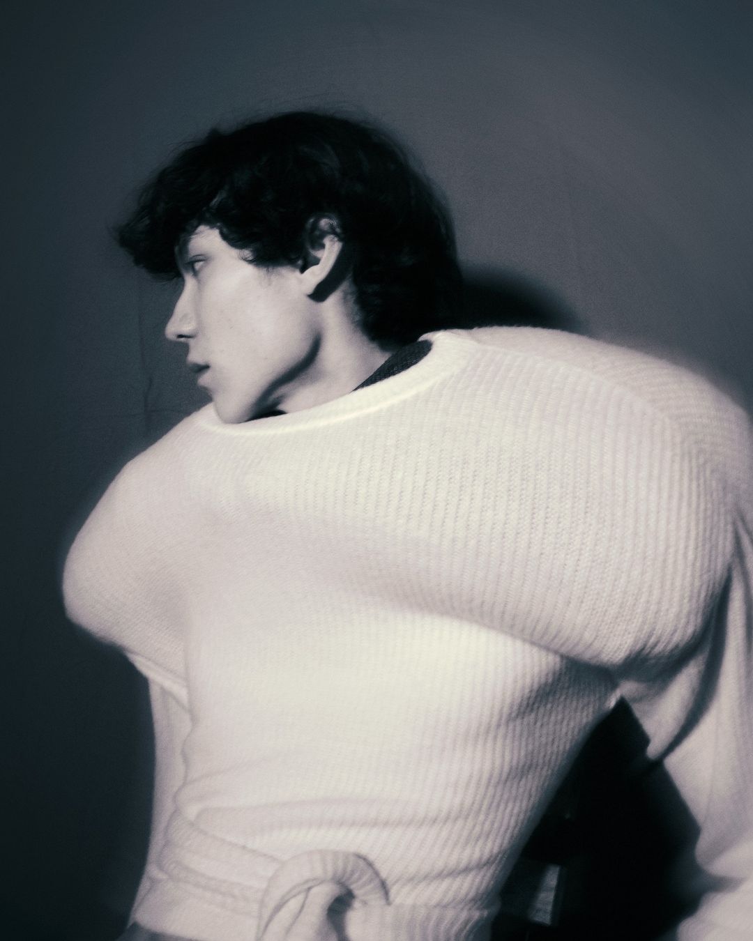 Monochromatic portrait of a young adult with dark hair wearing a white sweater, looking left, in a studio setting.