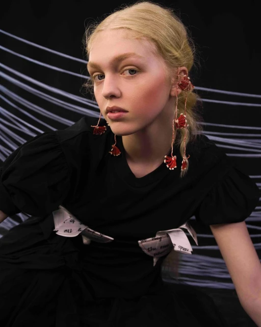 Young woman in a black dress with statement earrings and a paper-embellished belt poses in front of a black background with white lines.