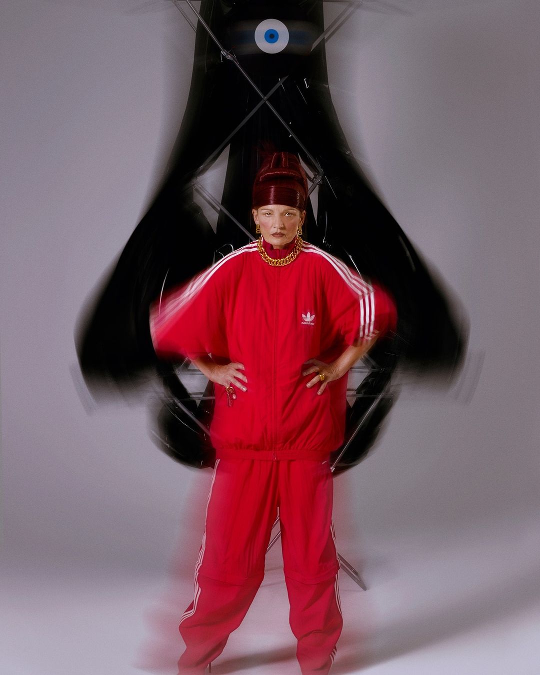 Woman in a red Adidas tracksuit stands in a photo studio with a large, abstract eye-shaped black sculpture and bright studio lighting.