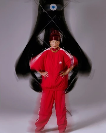 Woman in a red Adidas tracksuit stands in a photo studio with a large, abstract eye-shaped black sculpture and bright studio lighting.