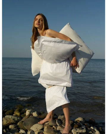 Woman holding white pillows standing on a rocky beach by the ocean under a clear sky by photographer Osman Özel.