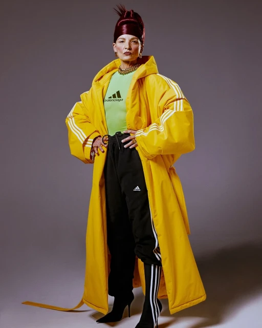 A stylish adult woman stands in a studio wearing a bright yellow Adidas robe, black track pants, and pointed heeled boots.