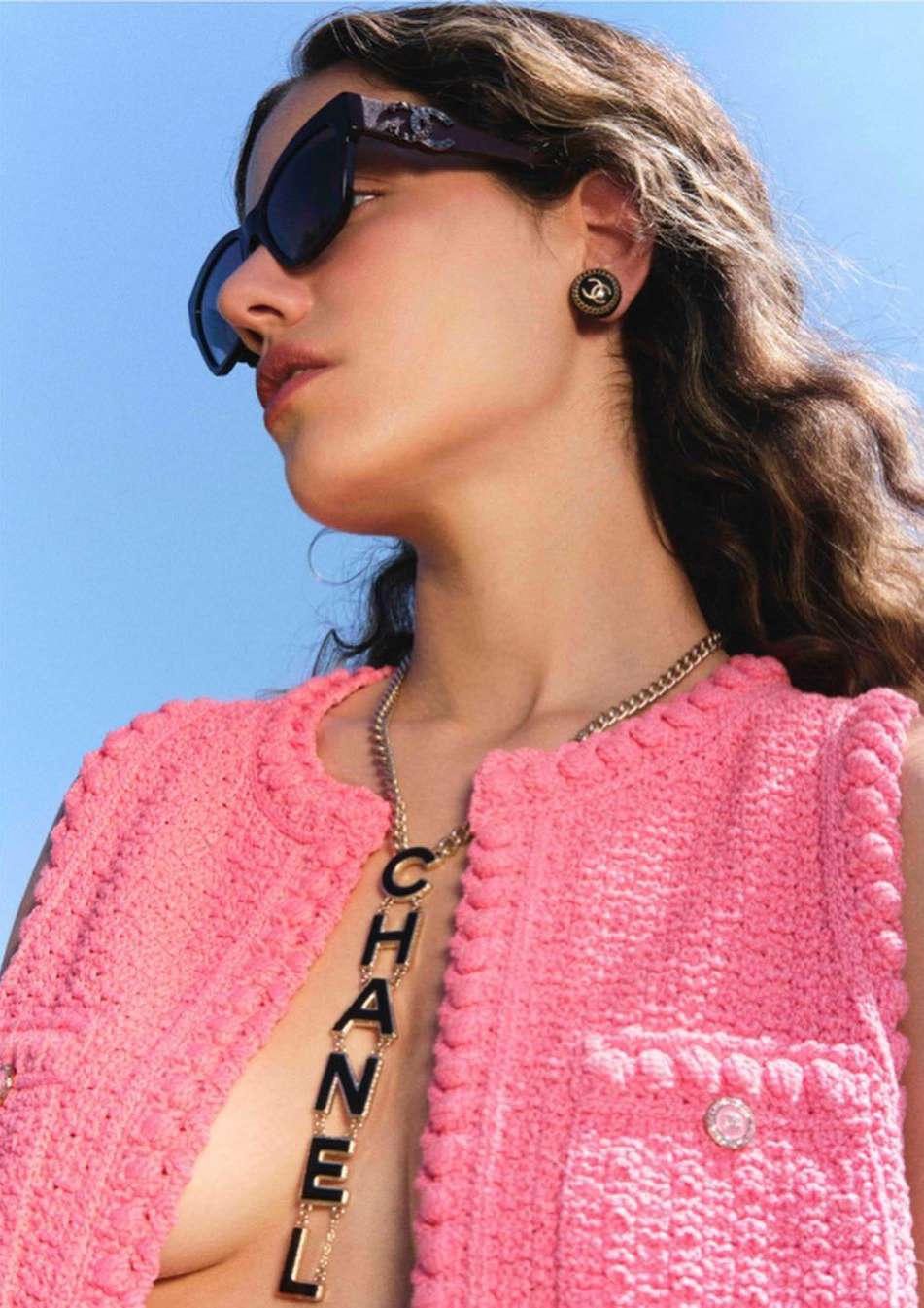 A woman wearing a pink textured tweed vest, black sunglasses, gold chain necklace and earrings against a clear blue sky background.