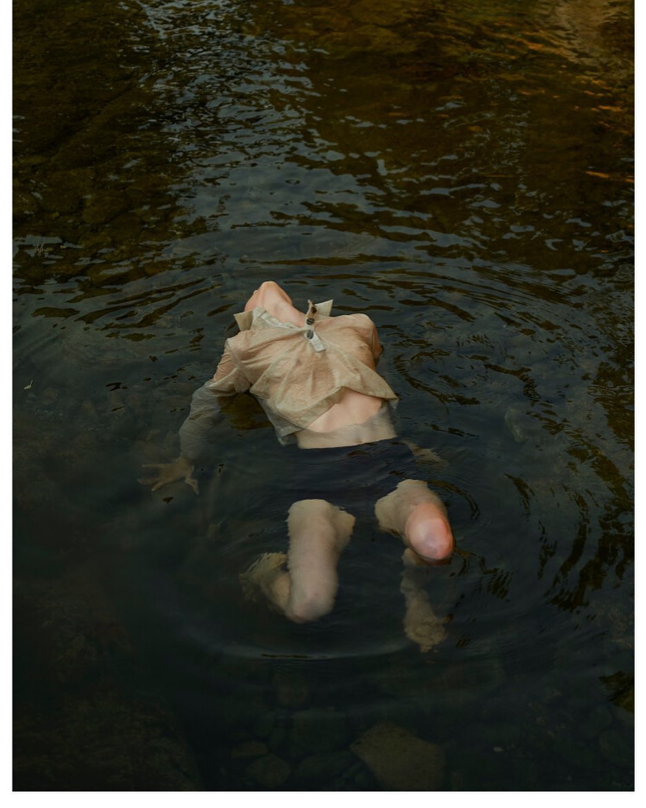 Person in a flowing shirt and black shorts immersed in a shallow, rocky river, capturing a sense of natural immersion and tranquility.