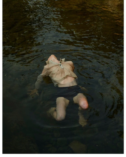 Person in a flowing shirt and black shorts immersed in a shallow, rocky river, capturing a sense of natural immersion and tranquility.
