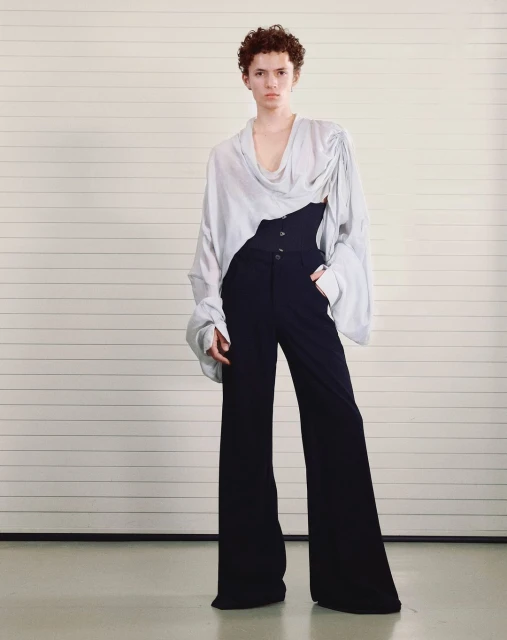 Full-length fashion shot of a model in a light-colored blouse with an asymmetrical neckline and dark, wide-legged trousers against a minimalist background.