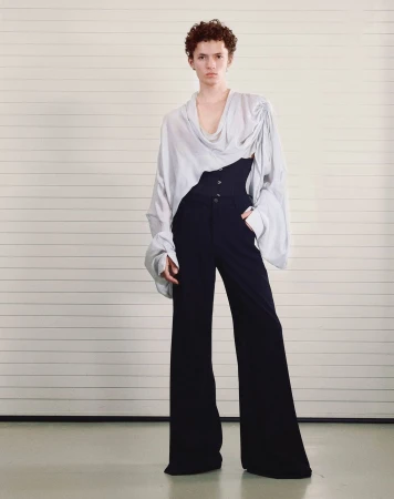 Full-length fashion shot of a model in a light-colored blouse with an asymmetrical neckline and dark, wide-legged trousers against a minimalist background.