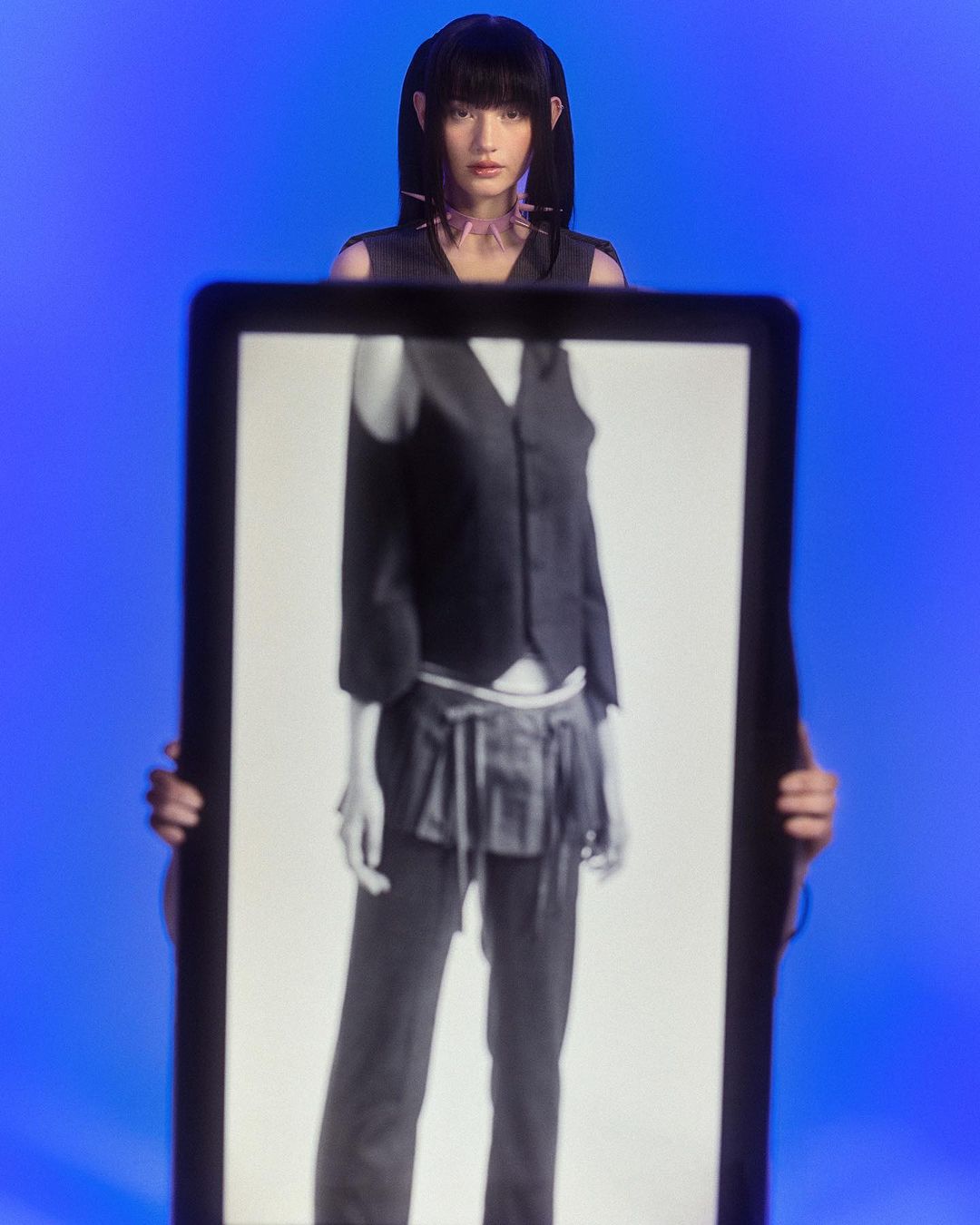 Portrait of an East Asian woman with blunt bangs and pigtails posing, while a person holds up a framed photo of a person in black and white.