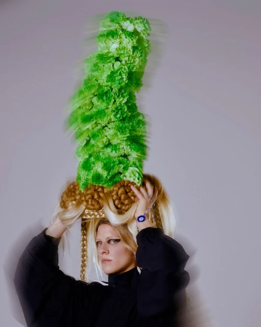 Woman with blonde hair, black top, and unique green headdress against a grey backdrop, showcasing a blend of fashion and artistic flair.