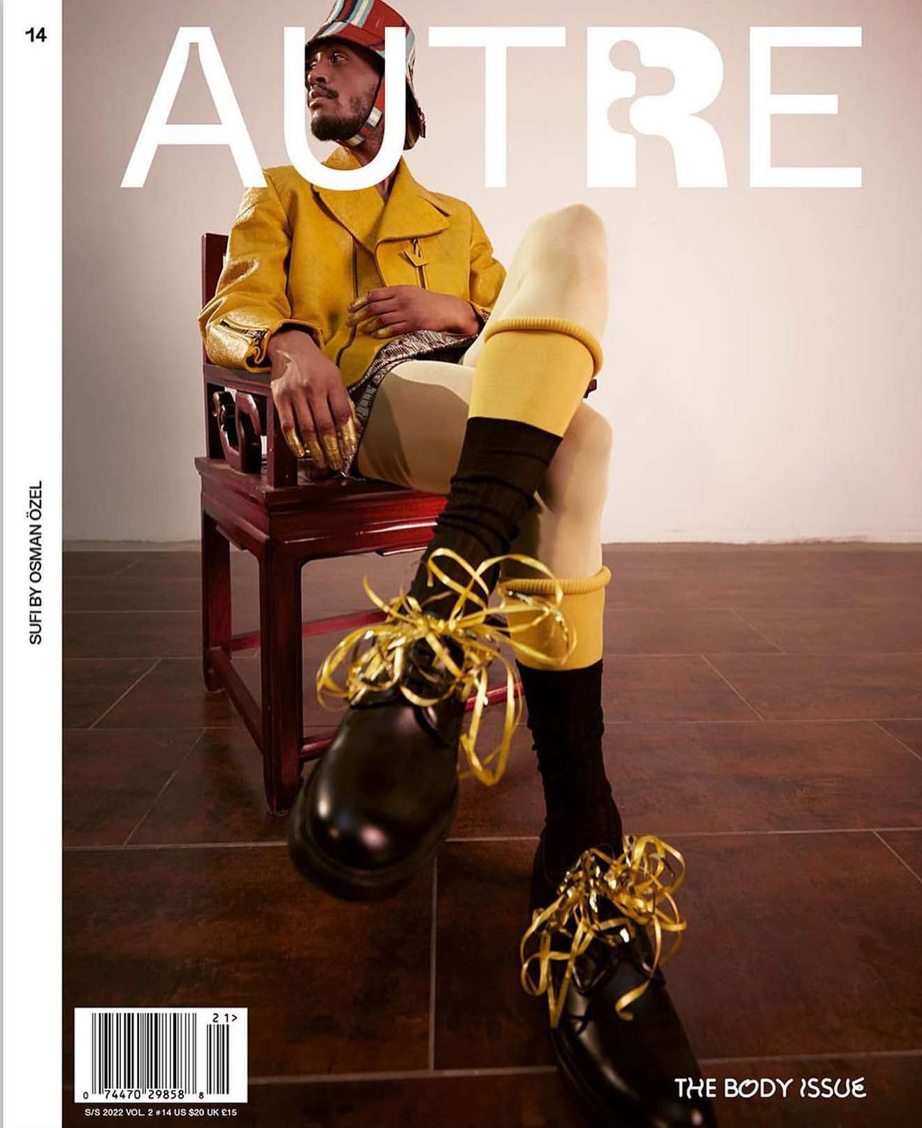 Magazine cover featuring a stylish adult male seated on an ornate chair with ribbons on his shoes, wearing a striking yellow outfit and helmet.