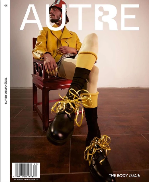 Magazine cover featuring a stylish adult male seated on an ornate chair with ribbons on his shoes, wearing a striking yellow outfit and helmet.