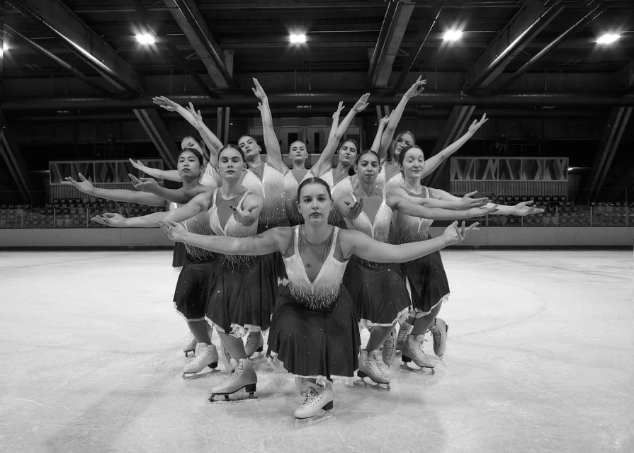 Synchronized ice skating team performing on ice rink. Female athletes in matching dresses with arms raised, teamwork and precision in monochrome.