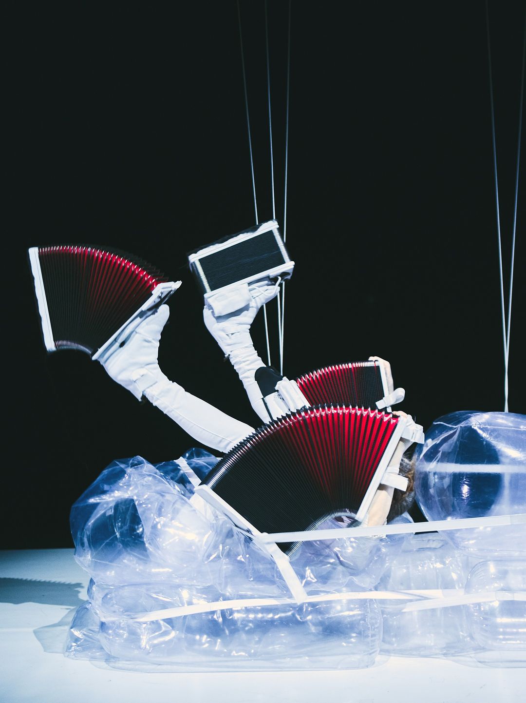 Surreal still life with accordion-like objects manipulated by gloved hands and suspended by wires, arranged on crumpled clear plastic against black.