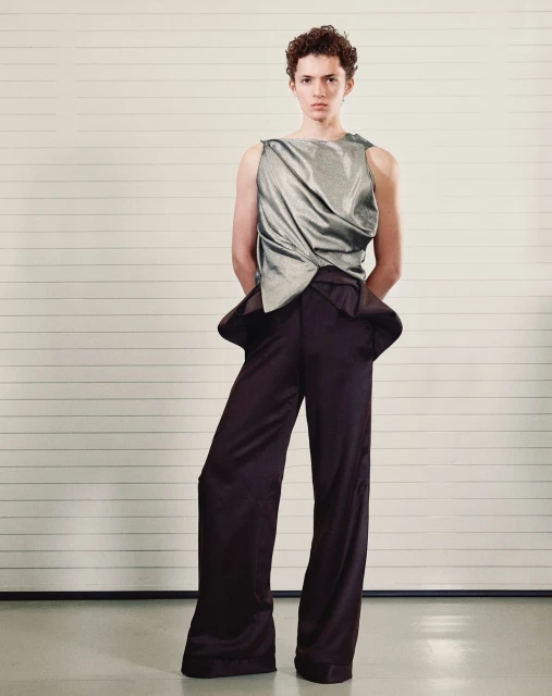 Young adult poses in silver draped tank top and wide-leg brown trousers against a studio background in a full length fashion portrait.