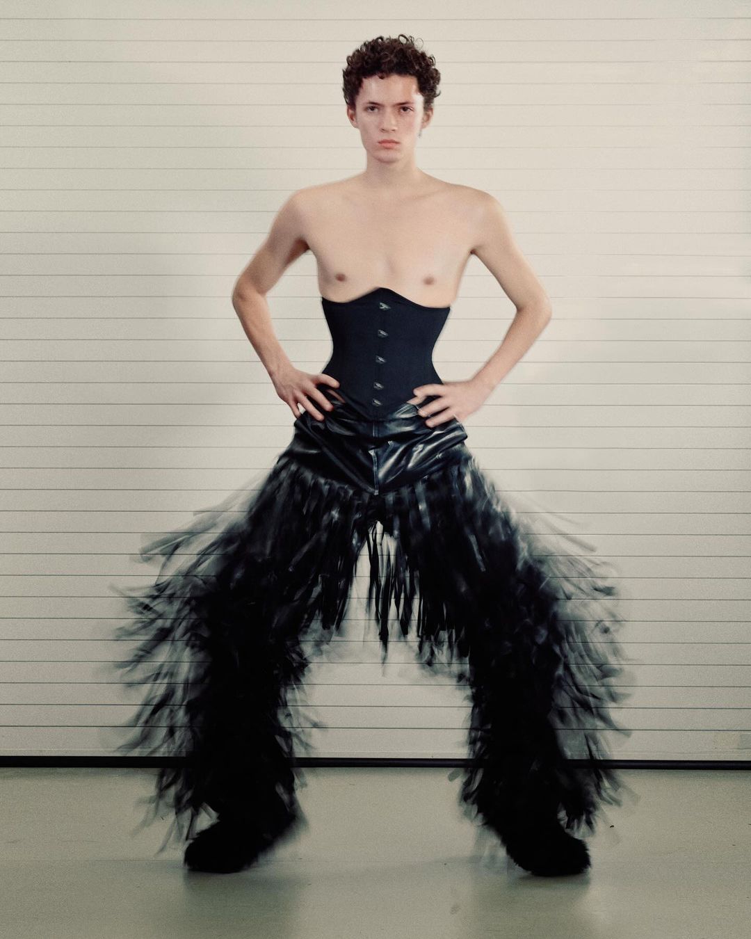 A person poses in a corset and dramatic black fringed trousers against a white garage door backdrop in a fashion forward portrait.