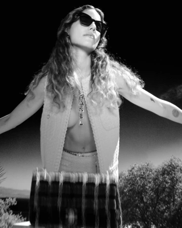 A woman with sunglasses wears a Chanel necklace, vest, and skirt outdoors against a dark sky and trees in a fashion-forward, black and white portrait.