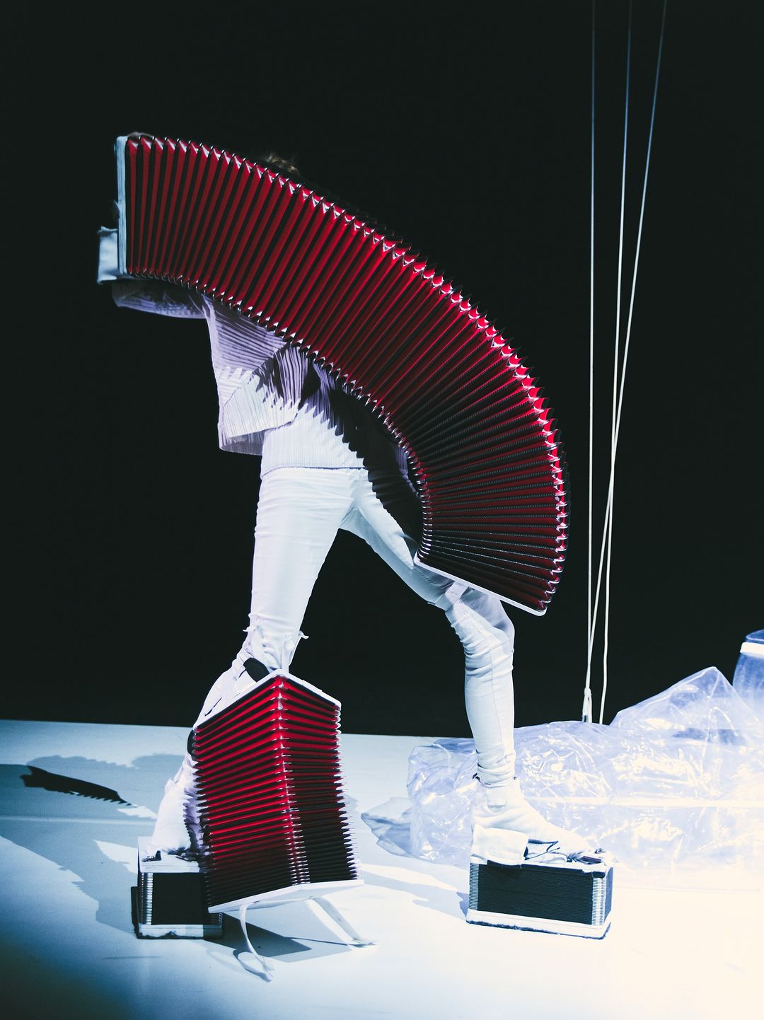Person in white outfit on a white platform with large red accordion-like apparatus, against a dark background in a studio shot.