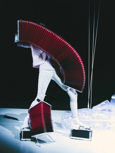 Person in white outfit on a white platform with large red accordion-like apparatus, against a dark background in a studio shot.