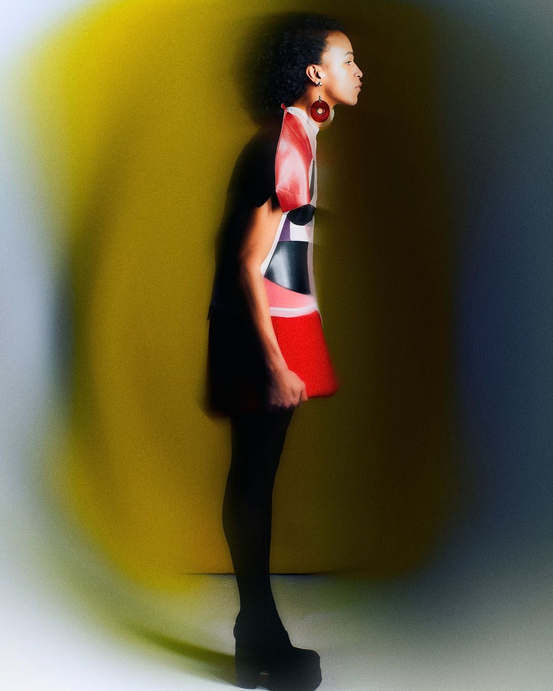 Side profile of a woman in a color-blocked mini dress with platform shoes posing against a gradient yellow and grey background in studio.
