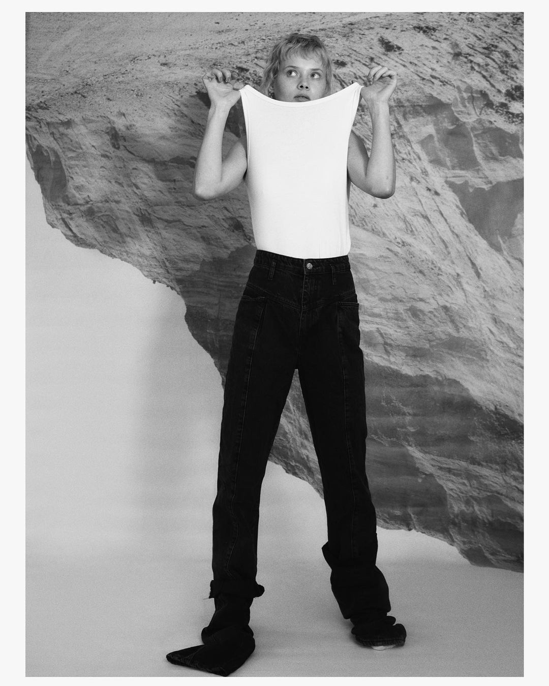 Young woman in casual style poses with a stretched white top against a black and white landscape backdrop, creating a studio-style image.