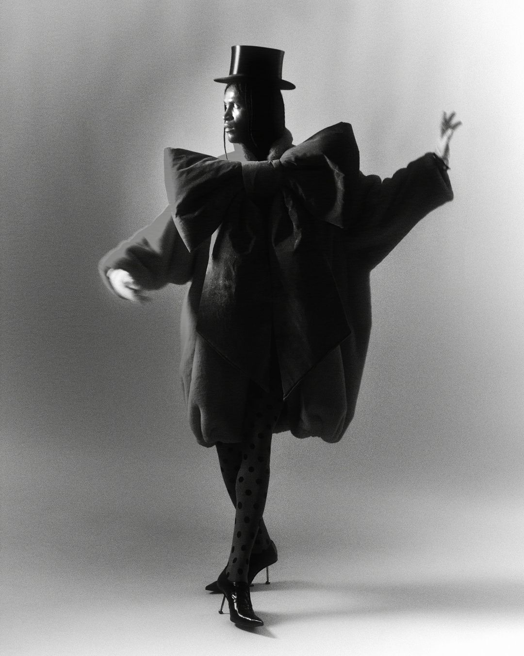 Fashion model in an oversized coat, polka dot stockings, and heels strikes a pose in a photo studio against a solid background.