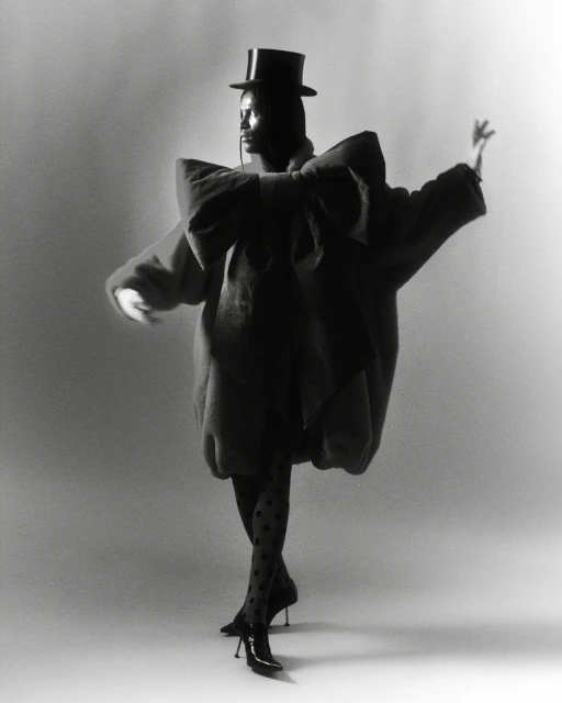 Fashion model in an oversized coat, polka dot stockings, and heels strikes a pose in a photo studio against a solid background.