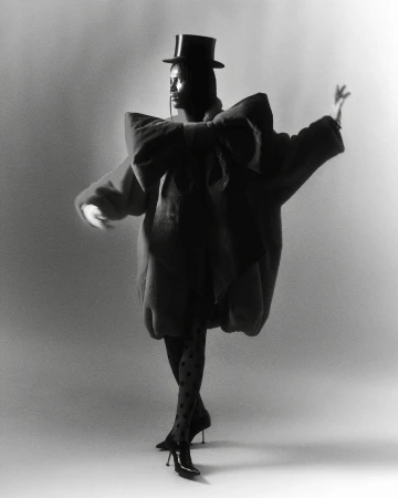 Fashion model in an oversized coat, polka dot stockings, and heels strikes a pose in a photo studio against a solid background.