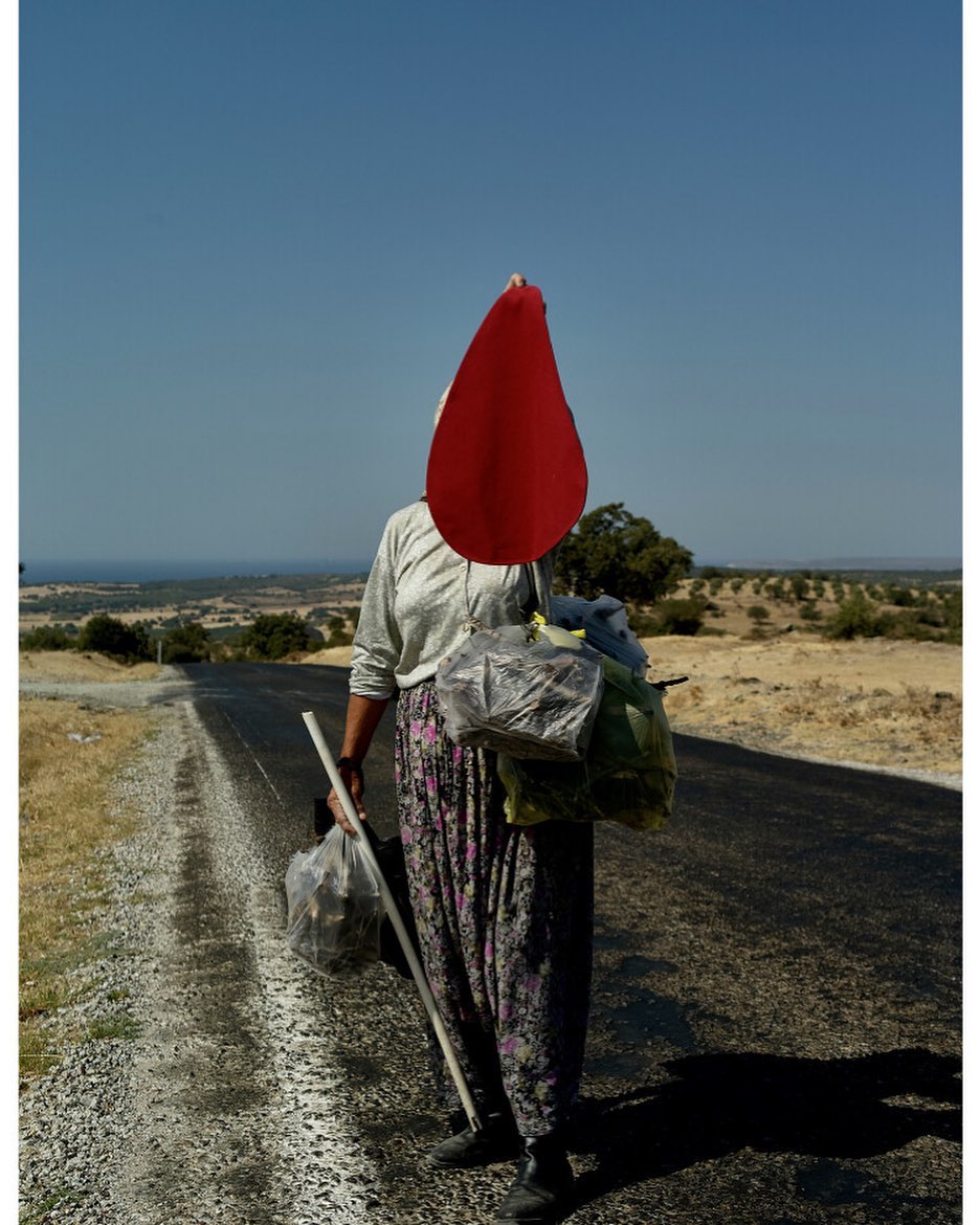 Woman with packages walks on road under blue sky on sunny day. Rural landscape and clear skies compose background. Walking stick in hand.