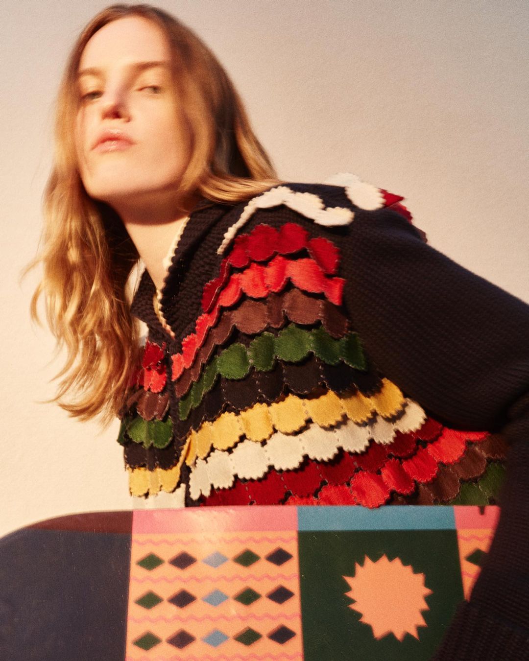 Woman in a multicolored garment with a pattern is posed indoors with a patterned object in a medium studio shot with natural lighting.