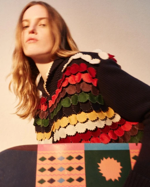 Woman in a multicolored garment with a pattern is posed indoors with a patterned object in a medium studio shot with natural lighting.