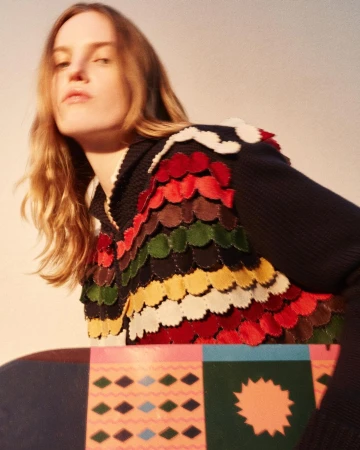 Woman in a multicolored garment with a pattern is posed indoors with a patterned object in a medium studio shot with natural lighting.