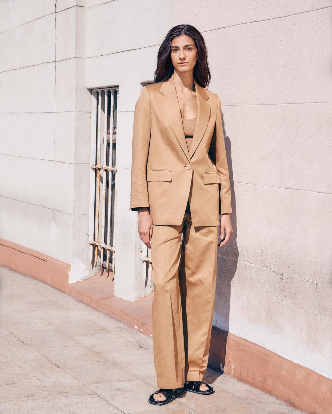 A brunette adult fashion model poses in a tan blazer and pants, paired with black sandals, against an exterior building wall.
