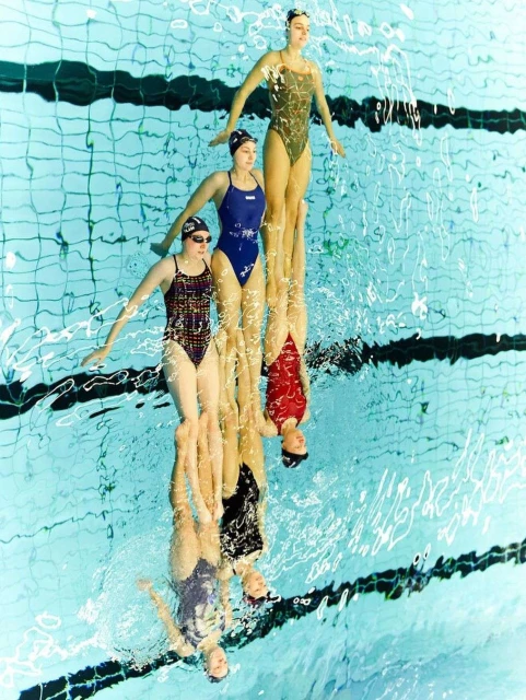 Synchronized swimming team performing in a pool with reflections. Three swimmers in formation wearing swimsuits and swim caps.