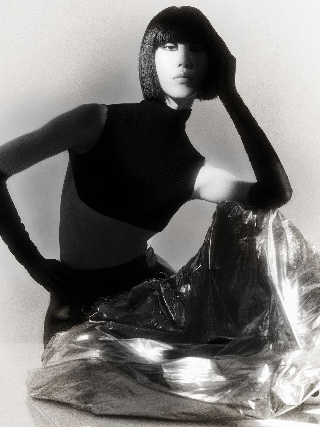 Monochrome portrait of an Asian woman with a bob haircut wearing a dark top and sitting in front of shiny silver fabric.