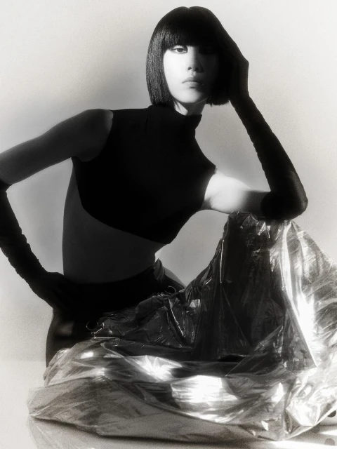 Monochrome portrait of an Asian woman with a bob haircut wearing a dark top and sitting in front of shiny silver fabric.