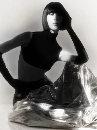 Monochrome portrait of an Asian woman with a bob haircut wearing a dark top and sitting in front of shiny silver fabric.