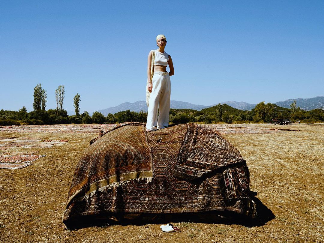 Fashion shot: a woman stands on a large rug in a field under a clear sky, wearing a matching white top and pants in a bright, outdoor setting.