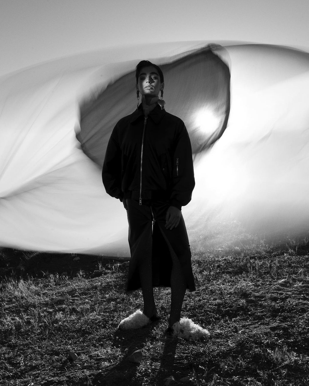 Black and white portrait of a woman standing in a field, wearing a jacket and skirt, with a sheet blowing behind her.
