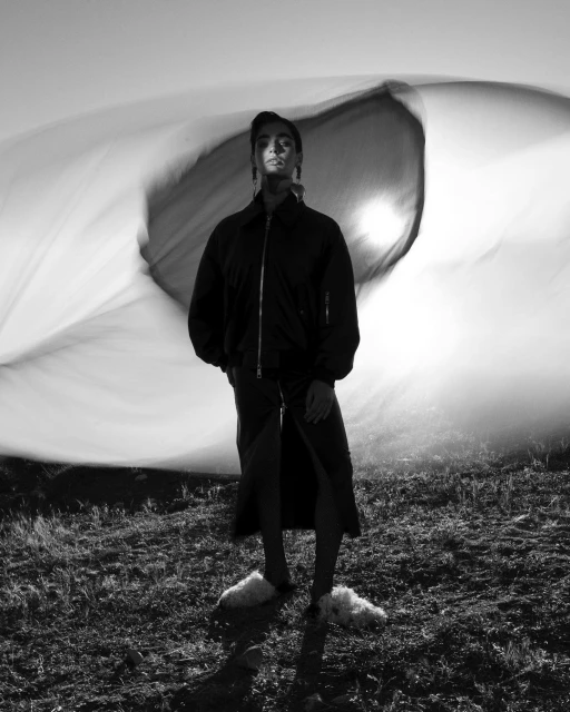 Black and white portrait of a woman standing in a field, wearing a jacket and skirt, with a sheet blowing behind her.