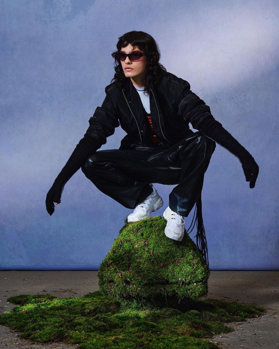 A young adult woman with sunglasses in a crouched pose atop a mossy rock, wearing a black jacket, gloves, and white sneakers.
