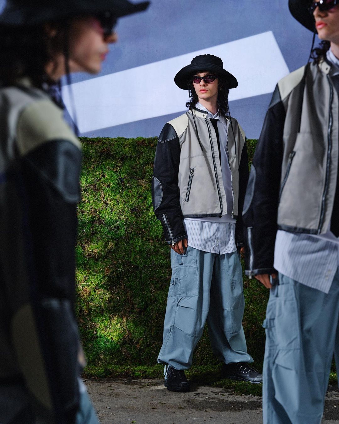 Three people in a line wear black hats, sunglasses, jackets with beige paneling, blue cargo pants, and black sneakers against a mossy wall.